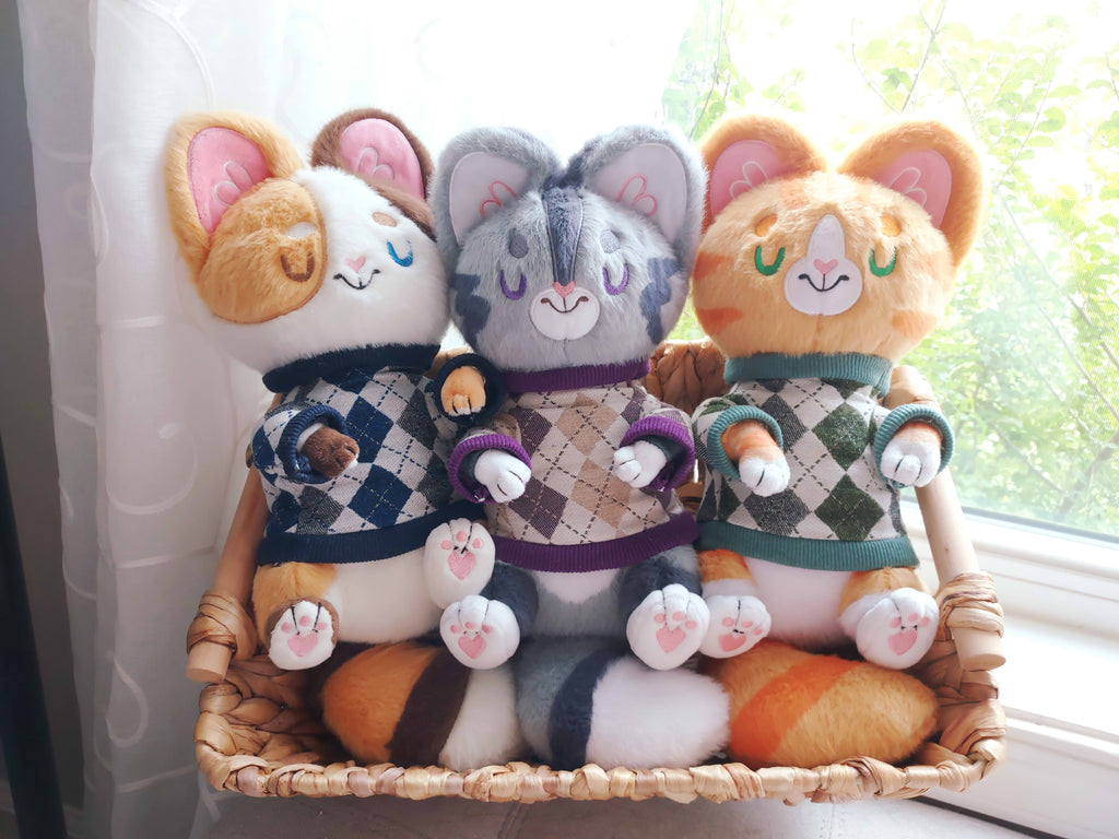 Snuggle Paws plush of Doodle the Calico cat, Domino the grey tabby, and Cheese the orange cat. They are wearing blue, purple and beige, and green argyle sweaters