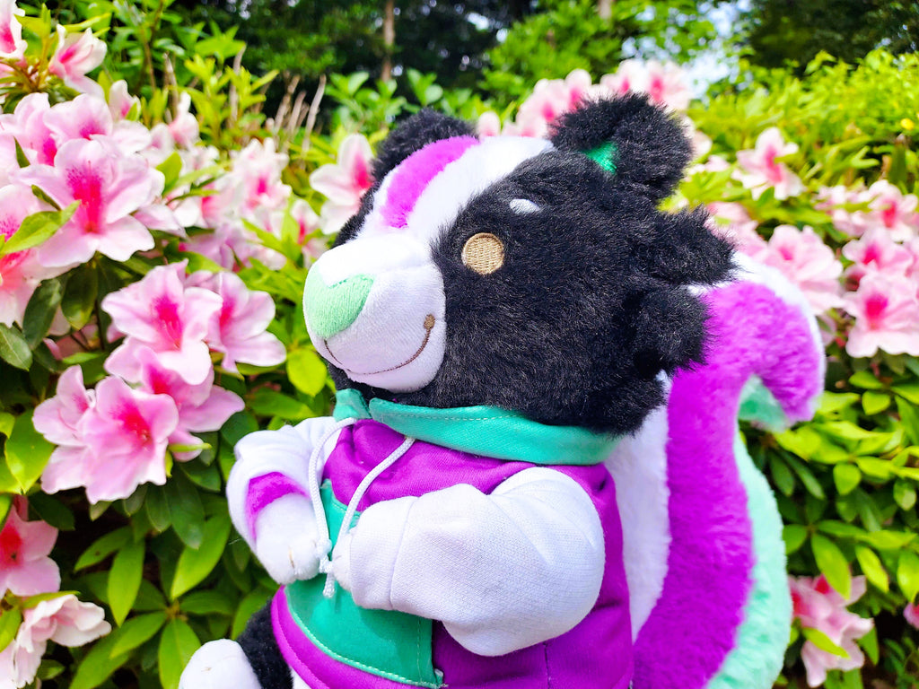 Plush of Pepper the skunk wearing a purple and green hoodie, sitting with pink flowers