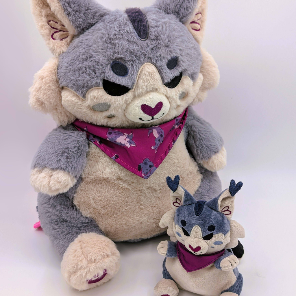 Plush backpack of a lynx wearing an aubergine bandana with a matching mini plush next to him.