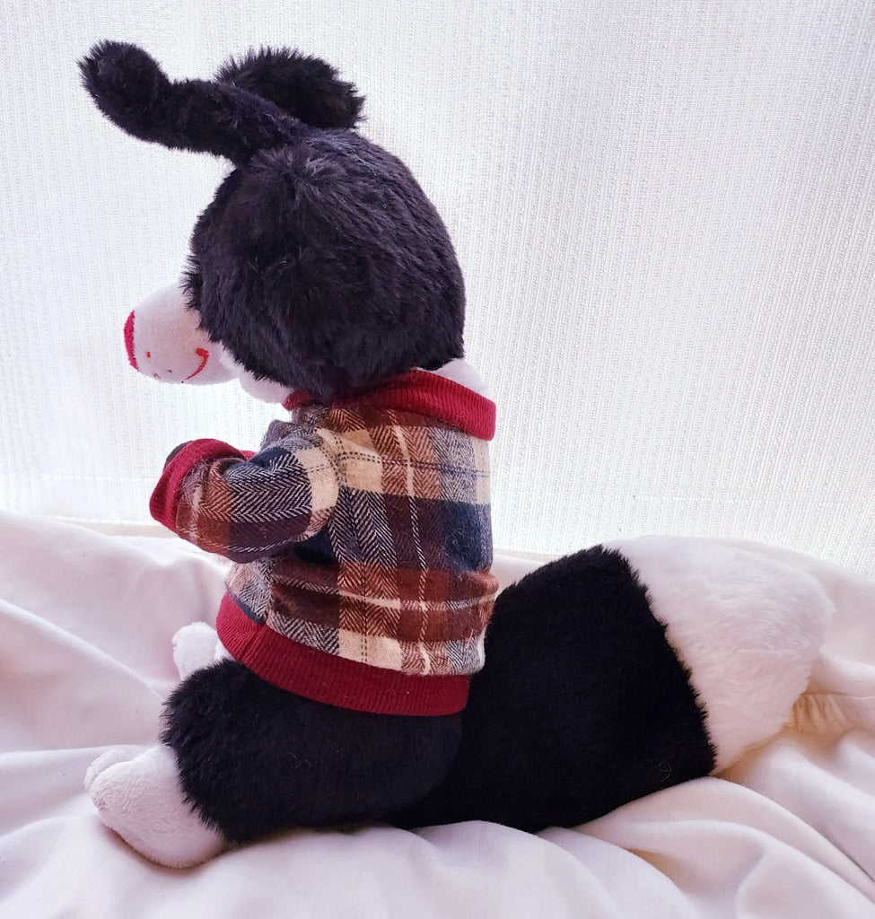 Side view of a plush toy of a black and white border collie with a red nose, wearing a red, beige, and blue plaid sweater