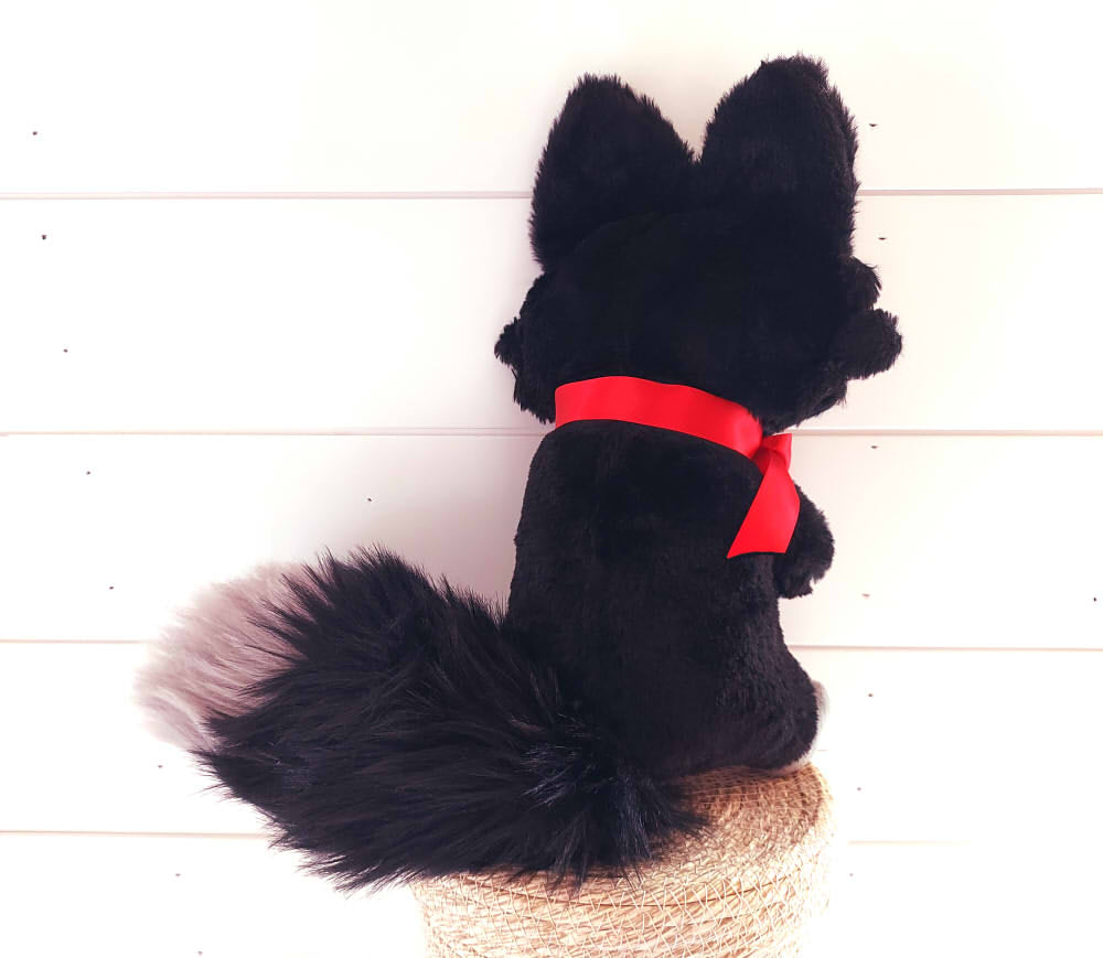 Back view of a plush toy of Crafty Tricks the fox who is black with red eyes and a charming red ribbon