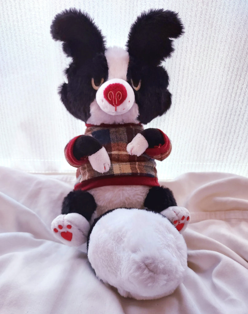 Plush toy of a black and white border collie with a red nose, wearing a red, beige, and blue plaid sweater