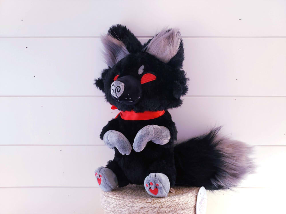 Plush toy of Crafty Tricks the fox who is black with red eyes and a charming red ribbon